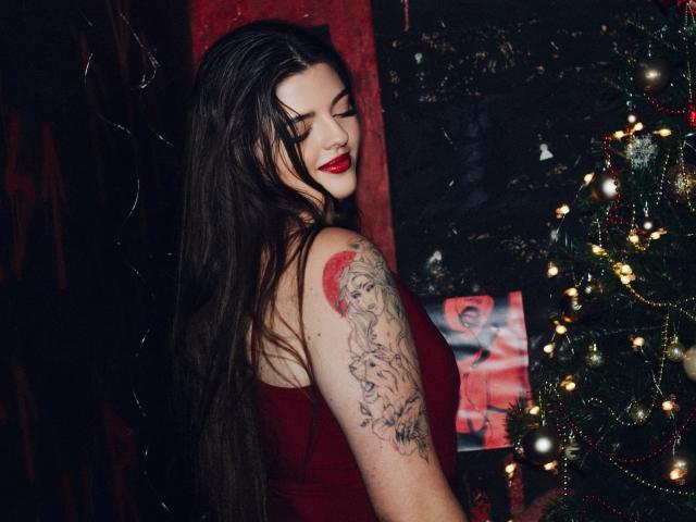 PamDares, com cabelos longos e escuros, batom vermelho e vestido vermelho, exibindo o braço tatuado, em pé ao lado da árvore de Natal, com os olhos fechados, aparentando ser sexy com um piercing no nariz. Seios visíveis..