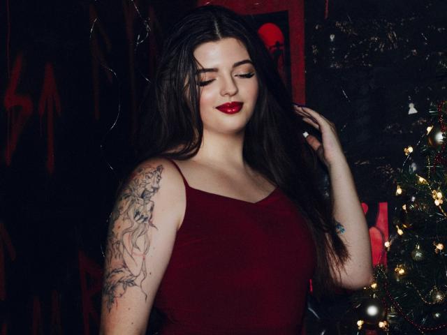 Pam, com cabelos castanhos longos, batom vermelho e um vestido vermelho, exibindo um braço tatuado, está em pé ao lado de uma árvore de Natal com luzes..
