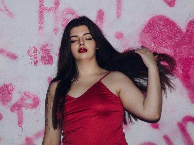 PamDares met lang donker haar, een rode satijnen topje, rode lippenstift, haar vasthoudend, bleke huid, naar beneden kijkend, een roze muur met graffiti op de achtergrond, zelfverzekerd, sexy, volop gevulde borsten..