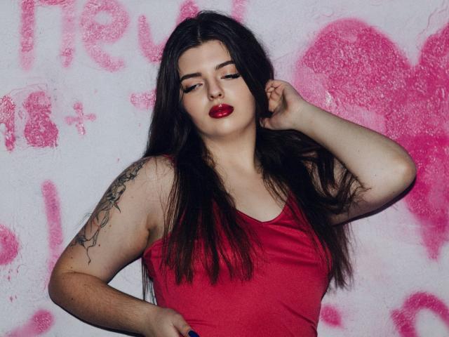 PamDares mit langem, schwarzem Haar, rotem Lippenstift und einem roten Tanktop, die ein Tattoo auf ihrem rechten Arm zeigt, mit der Hand im Haar, wirkt sie selbstbewusst. Helle Haut, volle Lippen..