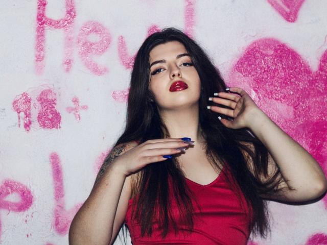 PamDares met lang donker haar, rode lippenstift, blauwe nagels, een tatoeage op de linkerarm, draagt een rode tanktop, haar hand raakt haar gezicht aan, ze kijkt omhoog, een witte muur met graffiti en roze vlekken..