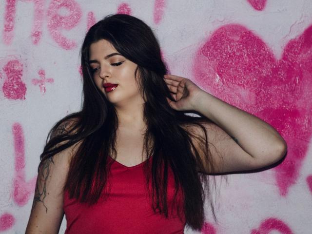 PamDares, con cabello largo y oscuro, lápiz labial rojo y camiseta sin mangas roja, con la mano en el cabello, tatuaje en el brazo izquierdo, piel pálida, sexy, segura de sí misma, mirando hacia abajo, fondo de grafiti rosa..