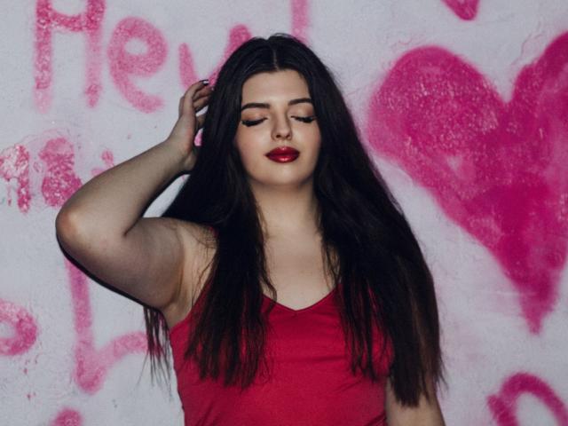 Pam, con lunghi capelli scuri, rossetto rosso e canotta rossa, occhi chiusi, mano tra i capelli, in piedi contro un muro bianco con graffiti rosa. Ha un bel sedere..
