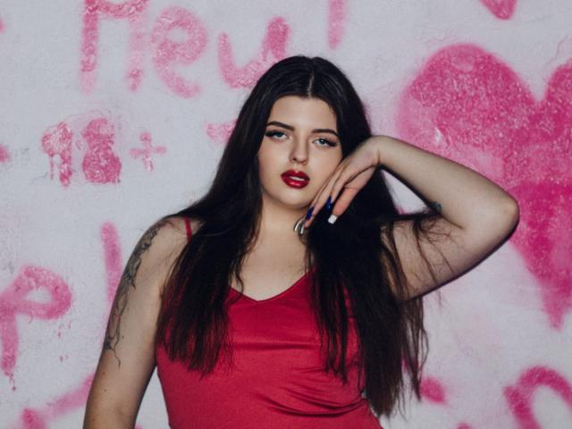 PamDares, met lang donker haar, rode lippenstift en een rood tanktopje, houdt een pen onder haar kin, heeft tatoeages op haar rechterarm en een zelfverzekerde blik..
