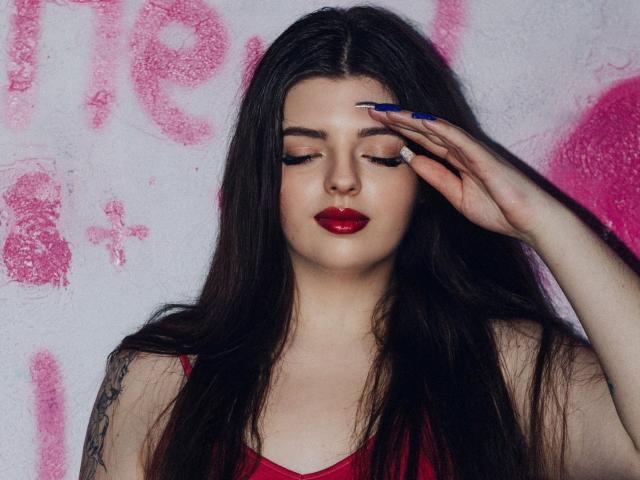 PamDares met lang donker haar, rode lippenstift en blauwe nagels, met gesloten ogen, haar hand op haar voorhoofd, gekleed in een rode top, met een tatoeage op haar linkerarm, op een witte muur als achtergrond..