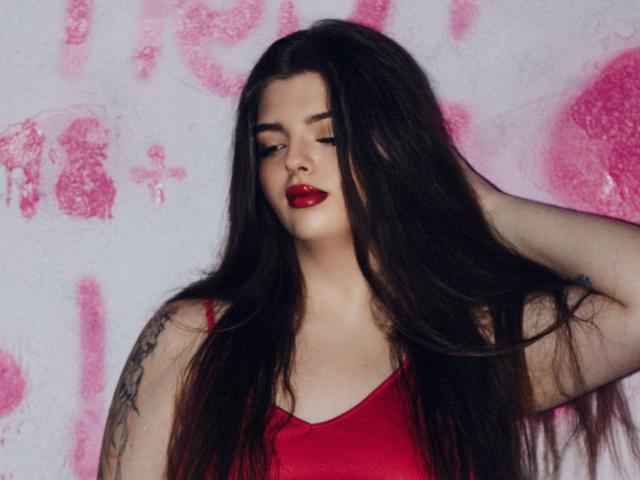 PamDares mit langen, dunklen Haaren, rotem Lippenstift, rotem Tanktop, Tattoo am Arm, Hand im Haar, nach unten schauend, rotem Make-up, weißem Hintergrund mit rosa Graffiti..