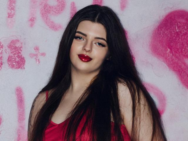 PamDares mit langem, schwarzem Haar, rotem Lippenstift und rotem Kleid, die leicht zur Seite schaut und ein leichtes Lächeln trägt, steht vor einer rosa Wand mit Graffiti..