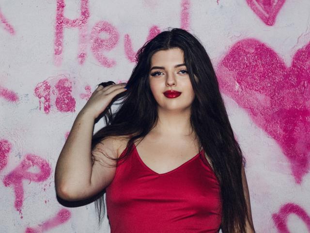 PamDares hat langes, dunkles Haar, roten Lippenstift und ein rotes Tanktop. Sie steht mit einer Hand im Haar und lächelt leicht in die Kamera..