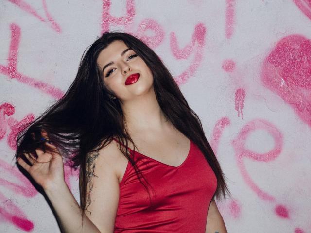 PamDares mit langem, dunklem Haar, rotem Lippenstift und einem roten Tanktop, wobei ein Tattoo auf ihrem rechten Arm sichtbar ist, hält sie ihre Haare. Ihre Brüste sind leicht sichtbar..