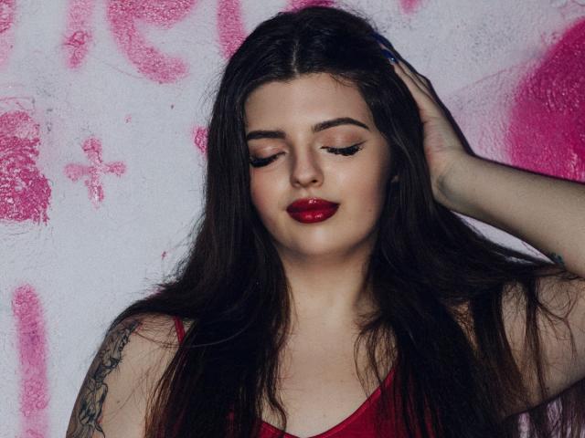 PamDares heeft lang donker haar, rode lippenstift en raakt haar hoofd aan met een hand. Ze draagt een rode top en heeft een tatoeage op haar rechterarm..
