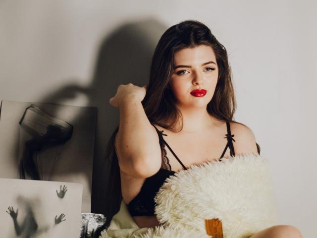 PamDares with red lipstick, black lace bra, and long brown hair, holding her hair up, sitting on a fluffy white pillow, looking at the camera with a serious expression.