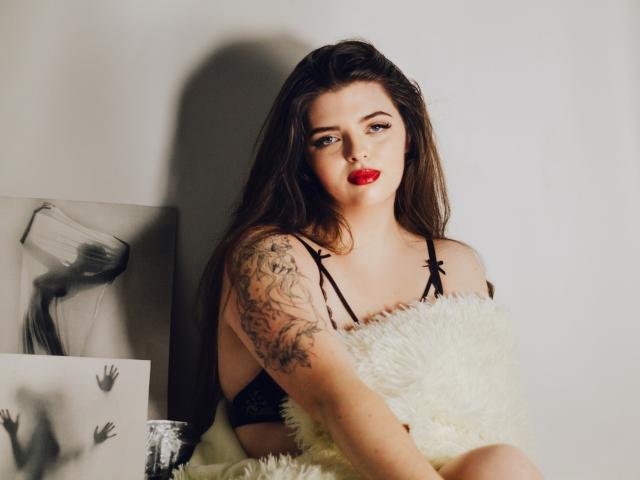 Pam, com batom vermelho, sutiã de renda preta e tatuagem no braço, sentada em um travesseiro branco fofo, segurando-o, olhando para a câmera, cabelo castanho, parede branca, arte em preto e branco..