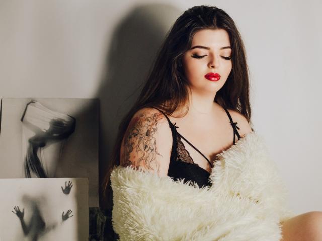 PamDares met rode lippenstift, donkere wenkbrauwen en lang bruin haar, draagt zwarte kanten lingerie en is gehuld in een pluizige witte jas, waarbij een tatoeage op haar rechterarm zichtbaar is..