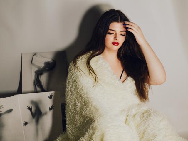 PamDares, com cabelos castanhos longos, batom vermelho e unhas azuis, vestindo um vestido branco fofo, com a mão no cabelo, sentada em frente a uma obra de arte em preto e branco à esquerda..