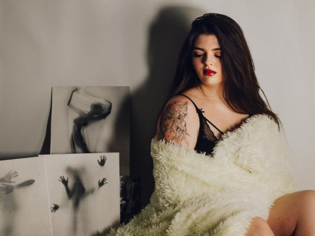 PamDares com lábios vermelhos, cabelos castanhos longos, sutiã de renda preta, casaco branco fofo e braço tatuado, sentada com uma expressão relaxada e sedutora. Arte em preto e branco ao fundo..