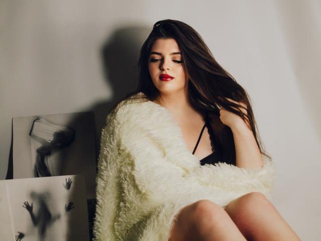 PamDares with long brown hair, red lipstick, black bra, and white fluffy sweater, sitting, looking down, holding hair, sexy, cute, soft lighting, shadow on wall, paintings in background.