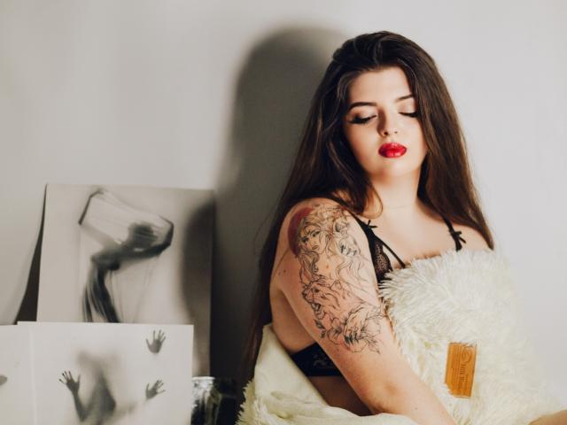 PamDares with long brown hair, red lipstick, black lace bra, tattooed arm, holding white fluffy pillow, eyes closed, sexy. Boobs visible, slightly covered by pillow.