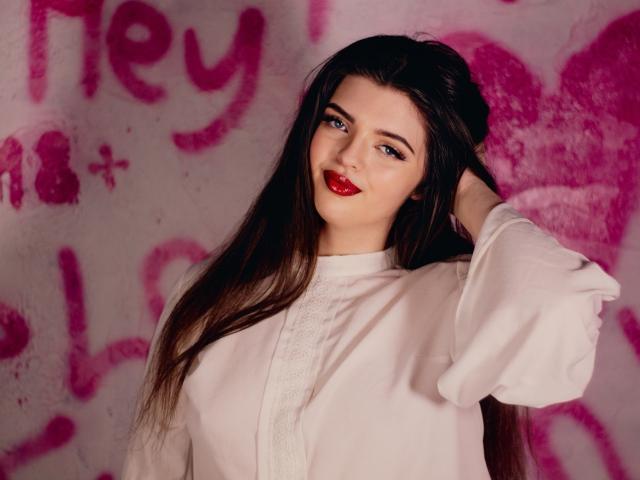 PamDares mit langen, dunklen Haaren, die ein weißes Hemd trägt, roten Lippenstift hat und blaue Augen, mit einer Hand in den Haaren, blickt in die Kamera und zeigt ihre Brüste leicht. Im Hintergrund ist Graffiti in Rosa zu sehen..