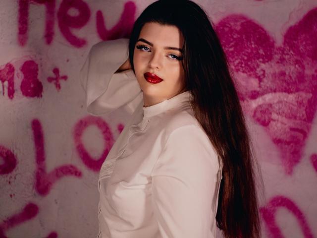 PamDares with long dark hair, red lipstick, white shirt, and confident smile, hand in hair, sexy, cute, and attractive, looking at camera.