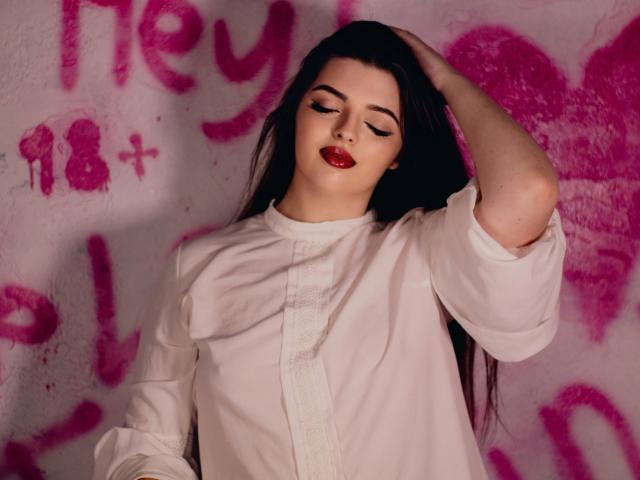 PamDares, con lunghi capelli scuri, indossa una camicia bianca, ha una mano tra i capelli, gli occhi chiusi, rossetto rosso, in piedi di fronte a graffiti rosa, appare sexy e sicura di sé..
