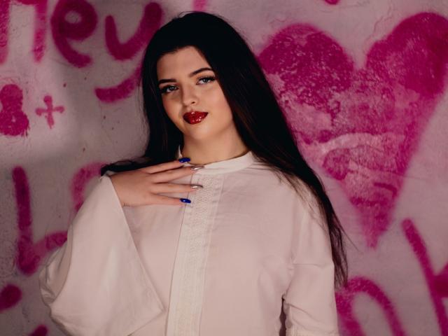 PamDares, con lunghi capelli scuri, rossetto rosso, unghie blu e un vestito bianco, con una mano sul petto, in piedi di fronte a un muro rosa ricoperto di graffiti. Il seno non è visibile..