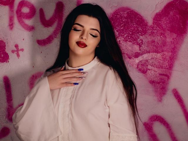 PamDares, con lunghi capelli scuri, rossetto rosso, smalto blu e vestito bianco, che si tocca il petto, in piedi di fronte a un muro rosa ricoperto di graffiti..