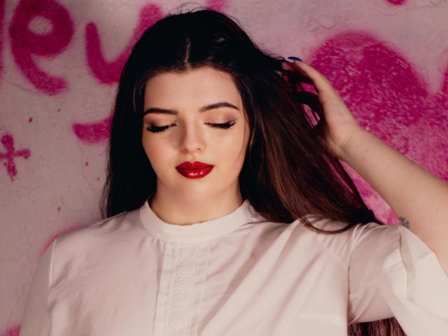 PamDares met lang bruin haar, rode lippenstift en gesloten ogen, gekleed in een witte blouse, met haar hand in haar haar, er sensueel uitzien, zacht licht, roze graffiti achtergrond, licht bleke huid..