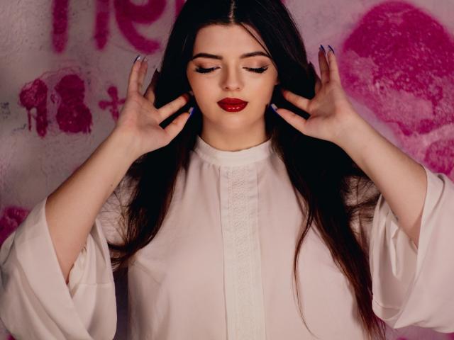 PamDares, com cabelos longos e escuros, batom vermelho e unhas azuis, vestindo um vestido branco, com as mãos nas orelhas, olhando para baixo, com um muro de grafite rosa ao fundo, iluminação suave..