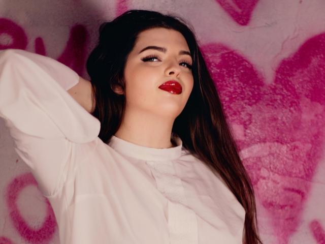PamDares con rossetto rosso, sopracciglia scure e lunghi capelli castani, indossa una camicia bianca, con un braccio alzato e lo sguardo rivolto verso l'alto. Sullo sfondo, ci sono graffiti rosa. Ha un'aria sexy e sicura di sé..