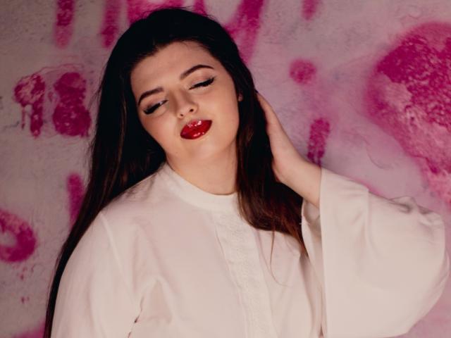 PamDares met lang bruin haar, rode lippenstift en een witte blouse, met haar in haar handen, ogen half gesloten, een roze achtergrond met spatten. Borsten licht zichtbaar onder de blouse. Iets rood aangelopen gezicht..