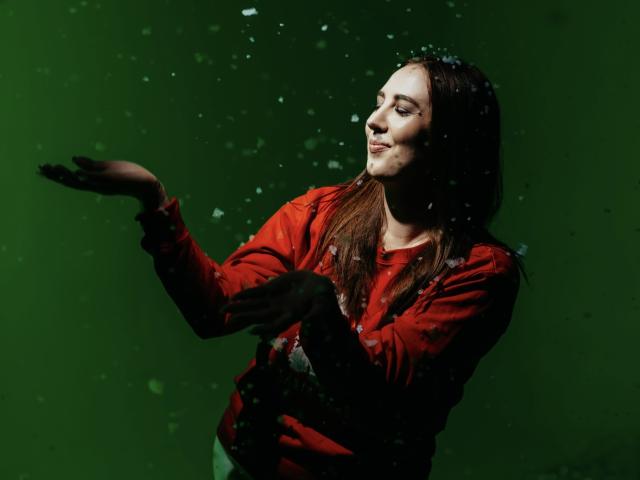 KaleyReed with long brown hair, red shirt, and green background, smiling while hands are up, snowflakes falling around her, eyes closed, soft light on her face.