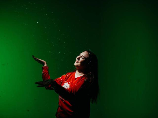 KaleyReed with long brown hair, wearing a red shirt, throwing black feathers, eyes closed, smiling, green background, feathers mid-air.