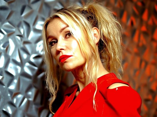 StarCrystal with blonde ponytail, red lipstick, and red dress, showing off her tits in a shiny, textured background with orange lighting. She looks confident and sexy.