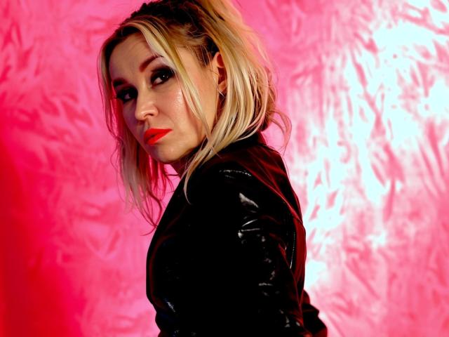 StarCrystal with blonde ponytail, black leather jacket, red lipstick, and smoky eye makeup, looking over shoulder, against pink, textured background. Her ass is slightly visible.