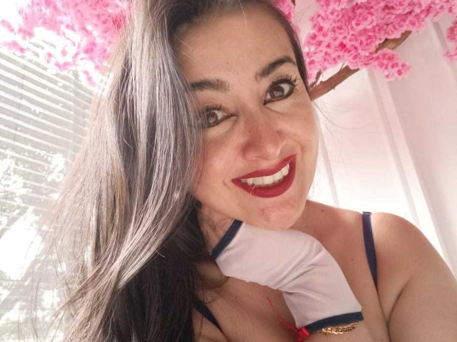 CherryHott69 with long black hair, red lipstick, smiling, wearing a blue-strapped top, and holding a white scarf, pink cherry blossoms in the background, and sunlight on her face.