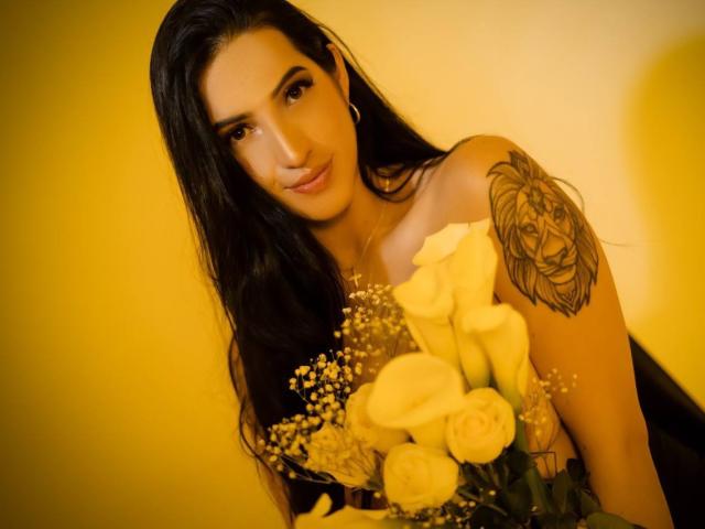 MeganBonnet with long black hair, holding a bouquet of yellow roses, has a detailed rose tattoo on her right shoulder, and a slight, sexy smile. Warm yellow light.