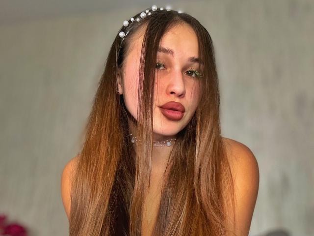 BrendaMoonx with long brown hair, wearing a pearl headband and choker, has pouty lips and green eyes, looking slightly to the side, showing her bare shoulders.