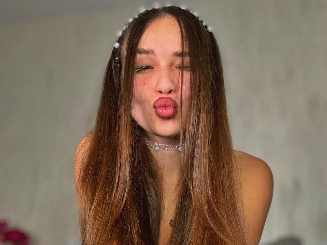 BrendaMoonx with long brown hair, pouty pink lips, and green eyes, wearing a delicate white headband and necklace, looking cute and playful. Boobs not visible.