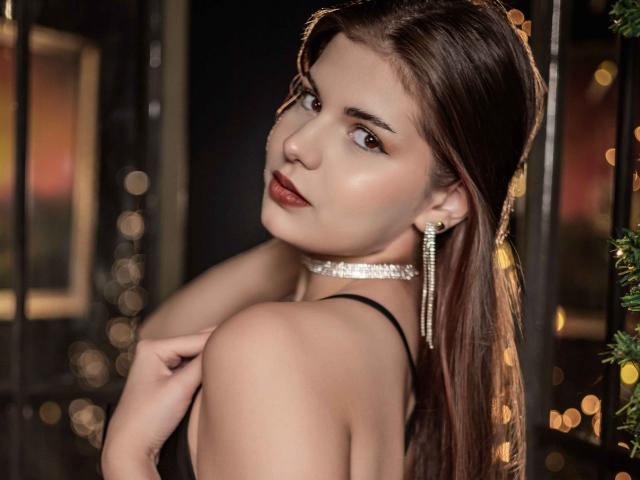 AdeliaLove with long brown hair, looking over her shoulder, wearing a diamond choker and dangly earrings, black dress, and red lipstick, boobs visible, sexy, soft light.