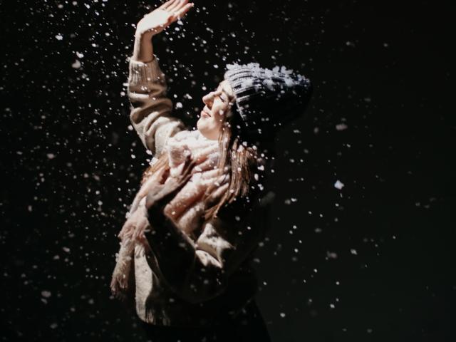 KaleyReed's tits are covered by a fluffy beige coat, black knit hat, and she's smiling up at falling snowflakes with her arms raised, dark background.