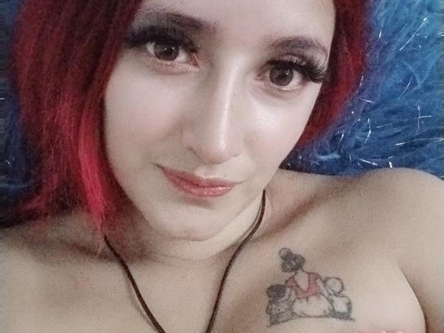 Hinnatha69, com cabelo ruivo, olhos castanhos e cílios longos, sorrindo para a câmera, mostrando uma tatuagem de uma mulher com um cachorro em sua mama direita..