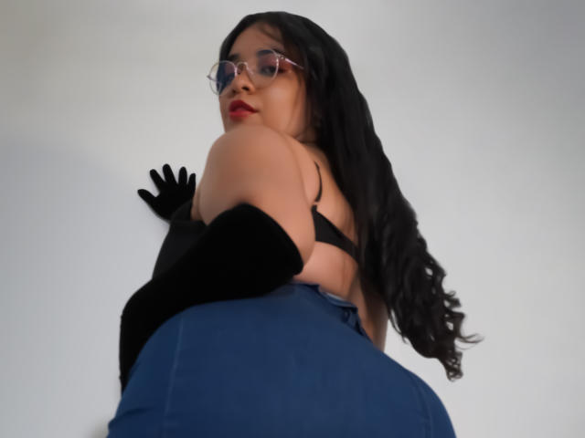 Alicia, con cabello negro largo, gafas, lápiz labial rojo, guantes negros, jeans azules y un sujetador negro, mostrando su gran trasero y pequeños senos, mirando por encima del hombro..