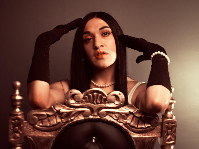 EmmaWalton with long black hair, orange lipstick, pearl necklace, and black gloves, posing with hands behind head, wearing white top, over ornate silver chair, showing small tits.