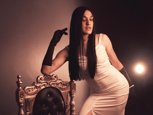 EmmaWalton with long black hair, wearing a white dress, black gloves, and pearl necklace, leaning on an ornate silver chair, eyes closed, showing off her ass in the shot.