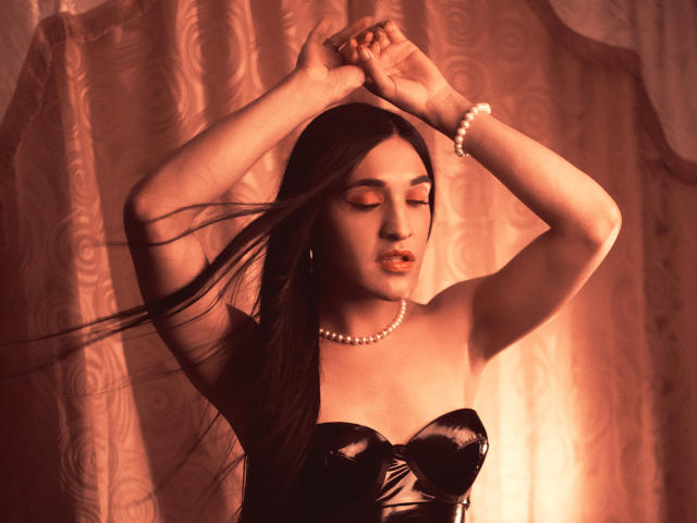 EmmaWalton with long dark hair, arms raised, wearing a black strapless top and pearl bracelet, eyes closed, lips slightly parted, soft red light, her tits are subtle.