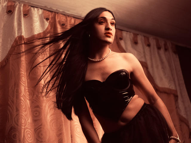 EmmaWalton with long dark hair, wearing a black strapless leather top, pearl necklace, and black skirt, her tits and ass highlighted, standing in a dimly lit room.