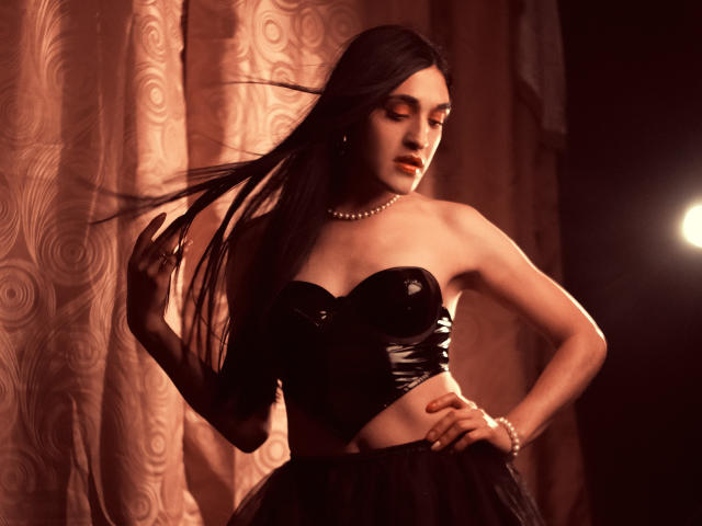 EmmaWalton with long black hair, wearing a shiny black strapless top, pearl necklace, and black skirt, holding her hair, standing in dim light, looking down with red lipstick.