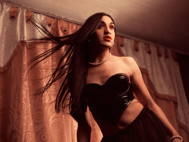 EmmaWalton with long black hair, wearing a black strapless leather top and black skirt, pearl necklace, and dark lipstick, her hair flying to the side.