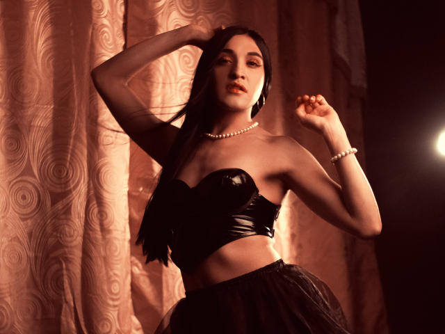 EmmaWalton with long black hair, wearing a black shiny strapless top and black skirt, pearl necklace, and bracelet, posing with arms raised, looking seductive, in dim lighting.