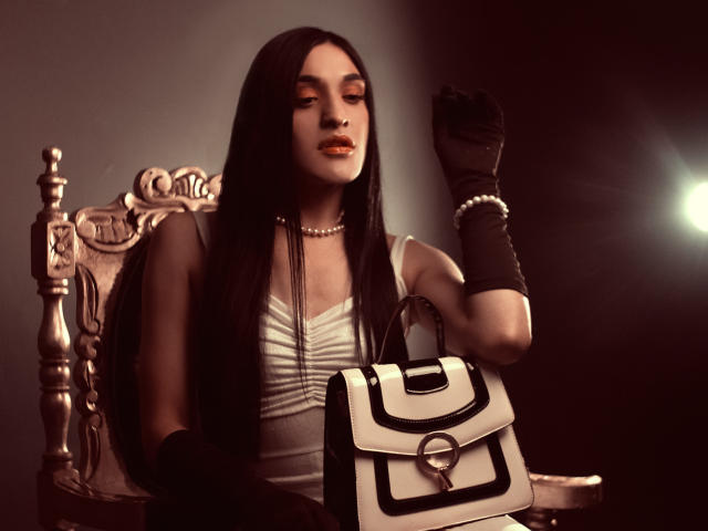 EmmaWalton with long black hair, orange lipstick, white dress, black gloves, pearl necklace, holding white and black handbag, sitting on ornate wooden chair, looking down.